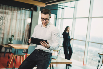 A young man in a white shirt stands in a spacious modern office and works on a tablet. Modern...