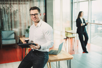 A young man in a white shirt stands in a spacious modern office with coffee and works on a tablet....