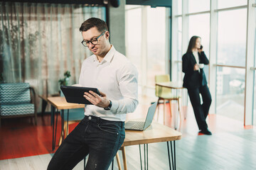 A young man in a white shirt stands in a spacious modern office and works on a tablet. Modern...