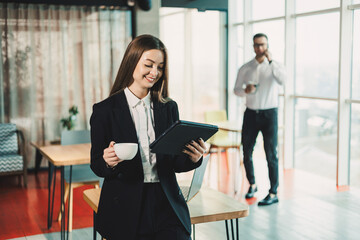 A young woman in a black classic suit is drinking coffee while standing in a modern office. Work in the office on a portable tablet.