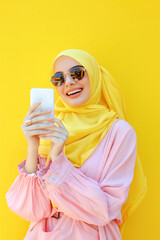 Obraz premium Cheerful Young Muslim Woman in Hijab Using Smartphone on Vibrant Yellow Background