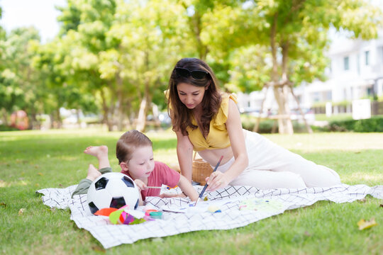 Middle-aged woman teaching a cute son Draw on paper using a cloth spread on the grass in the village park on a family day out while waiting for dad to go to work.
