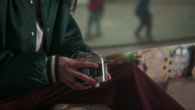 Tilt up shot of young gen Z girl sitting at skatepark, inserting cassette tape into player, putting on headphones and enjoying music
