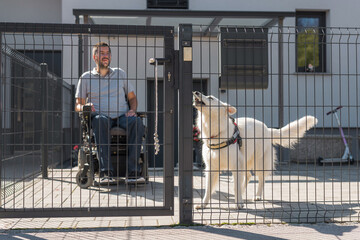 Man in a wheelchair leaving his house yard with the help of a service dog opening and closing the...