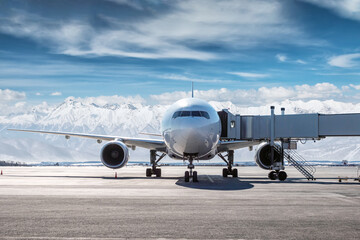 Front view of the white wide body passenger aircraft at the airbridge on the airport apron on the background of high snow capped mountains