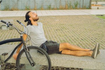 young latin man resting on a bench next to his bicycle, getting some sunshine