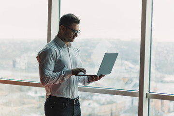 A young handsome man with a laptop stands on the background of a large panoramic window in the sky. A modern office with large windows and an office worker