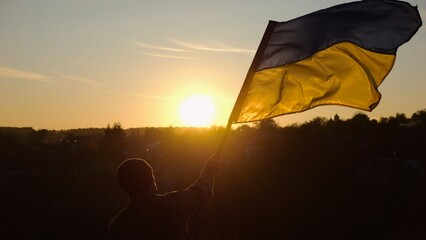 Young man in military uniform waving flag of Ukraine against beautiful sunset at background. Male ukrainian army soldier lifted national banner at countryside. Victory against russian aggression - 757492877