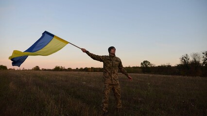 Young male military in uniform jogging with flag of Ukraine on meadow at dusk. Soldier of ukrainian army waving blue-yellow banner in honor of victory against russian aggression. End of war. Slow - 757493055