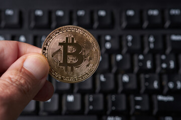 A hand is shown holding a bitcoin in front of a keyboard. The hand is positioned above the keyboard, with the bitcoin clearly visible. This image conveys a concept of digital currency and technology i