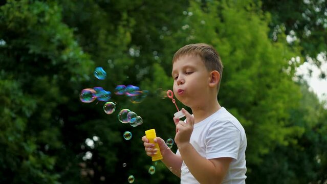 Preschooler boy spends time blowing soap bubbles creating playful atmosphere slow motion. Playing in summer city park cheerful boy happily blows bubbles. Young boy delights in blowing soap bubbles
