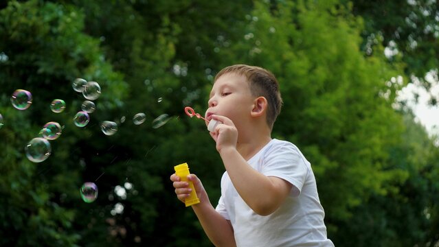 Preschooler boy spends time blowing soap bubbles creating playful atmosphere slow motion. Playing in summer city park cheerful boy happily blows bubbles. Young boy delights in blowing soap bubbles