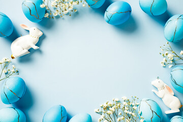 Frame of blue Easter eggs with rabbits and white flowers on pastel blue background.