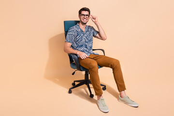 Full length photo of handsome businessman touching specs sitting comfortable boss chair enjoying office isolated on beige color background