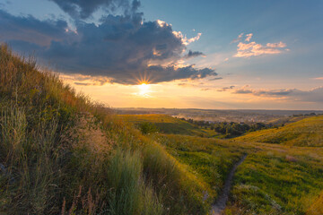 Naklejka na ściany i meble The majestic dusk scene with motley high grass and wildflowers covered hills in front of a beautiful sunset sky with clouds. Summer nature background.