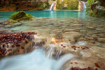 Calm waters cascade over rock formations, adorned with autumn leaves at the Urederra River, set against a backdrop of a serene waterfall