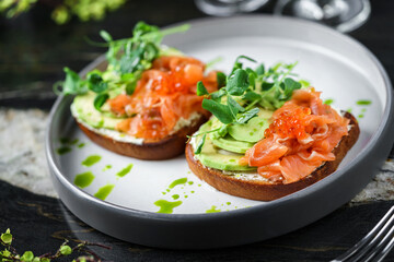 Sandwiches with cream cheese, avocado, slices salmon, red fish caviar and microgreens on plate over table with drinks. Healthy breakfast food, top view