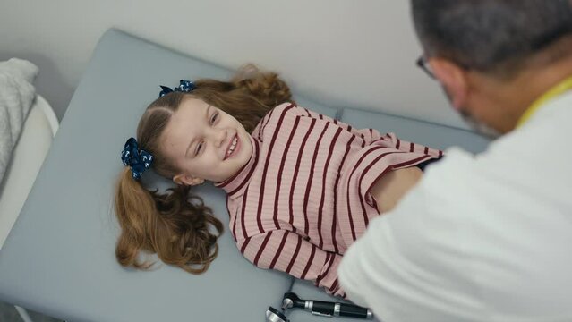 Over-the-shoulder shot. A little girl patient lies on her back on the medical examination couch and laughs while her abdomen is examined by a pediatrician. A male doctor palpation a child's abdomen
