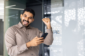 A cheerful man in a casual shirt celebrates success with a raised fist in an office setting,...