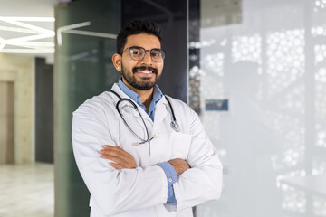 Smiling male doctor in white coat with stethoscope standing confidently in a hospital setting, arms crossed.