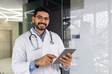 A cheerful male healthcare professional wearing glasses stands in a clinic, holding a digital tablet and radiating confidence.