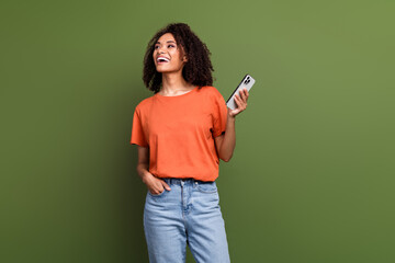 Photo of shiny dreamy woman wear orange t-shirt communicating modern gadget looking empty space isolated green color background