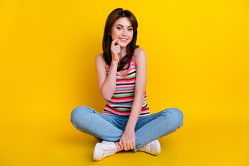 Full body photo of cheerful girl wear striped tank denim trousers sitting on floor finger on teeth isolated on yellow color background