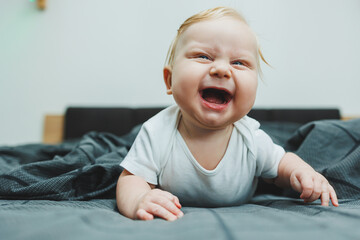 A cheerful baby boy with beautiful big eyes is lying on the bed at home in a white bodysuit on gray bedding. The child is lying in bed at home - Powered by Adobe