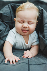 A cheerful baby boy with beautiful big eyes is lying on the bed at home in a white bodysuit on gray bedding. The child is lying in bed at home