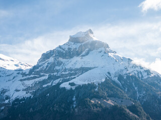 Fototapeta na wymiar Early morning or late afternoon light bathes a snowy Engelberg mountain peak, with rocky outcrops and forested slopes beneath a pale blue sky with wispy clouds.