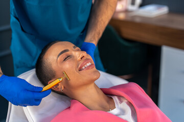 Wealthy woman getting massage with face golden roller massager at beauty clinic, sitting...