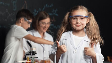 Smart caucasian girl showing thumb while teacher and student doing experiment behind. Child looking...