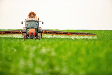 Deurstickers Tractor with sprayer arms extended applies treatment to crops on a vast, green farmland © oticki