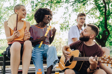 Multicultural friends sitting in city park and singing with guitar.