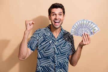 Photo portrait of young brunet hair guy raised fist up hold money to fly vacation lottery winner isolated on beige color background