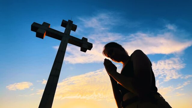 Silhouette of young woman is folding hands and praying against Georgian huge stone cross monument and blue sky at sunset - low angle. Christianity, Orthodoxy, hope, religion and spiritual concept