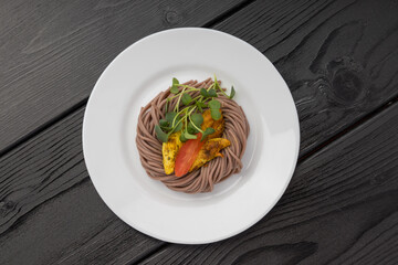 A plate of brown noodles topped with green leaves, yellow and red spices, dark wood table.