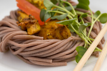 Close-up of brown noodles with chicken, tomato, and greens, white plate, focused textures.