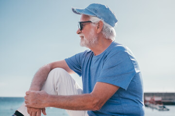 Smiling senior bearded man sitting outdoors face the sea enjoying sunny day, travel, vacation,...