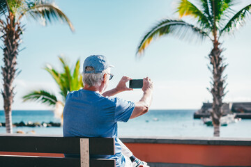 Back view of senior man using smartphone to take pictures sitting outdoors on bench face the sea enjoying sunny day, travel, vacation, retirement lifestyle