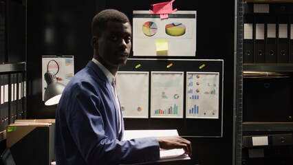 Portrait of african american investigator standing in evidence room, reading case files. In dimly...