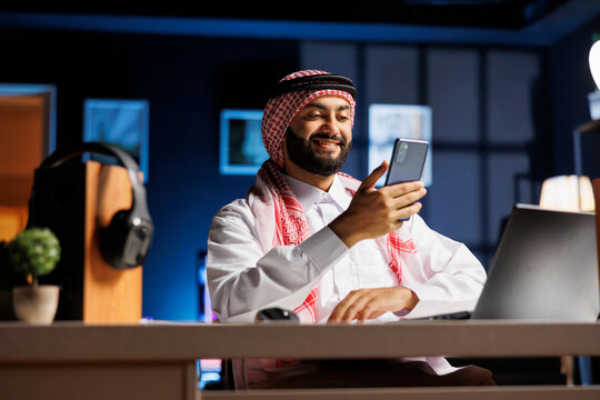 Enthusiastic Islamic man surfing the net on his digital mobile device. Detailed view of a male Muslim person with a smile, sitting at the table using his smartphone.