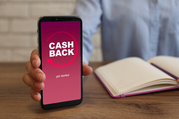 Woman holding smartphone with word Cashback at wooden table, closeup
