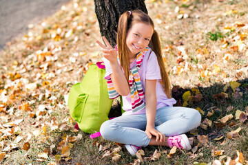 Photo portrait of ponytails cute small girl rucksack sit under tree show v-sign dressed stylish garment elementary school student park