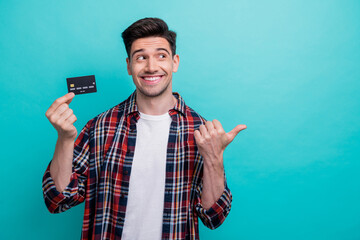 Photo of cheerful dreamy man dressed plaid shirt rising bank card pointing thumb empty space isolated turquoise blue color background