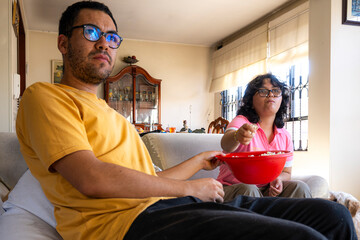 Siblings watching tv and eating popcorn from red bowl in apartment