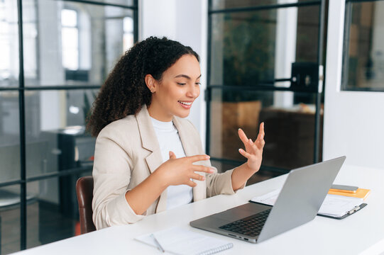 Online discussion. Positive latino or hispanic business woman, ceo, team leader, sits in modern office, looks at laptop screen, gesturing with hands, talking with colleagues by video conference, smile