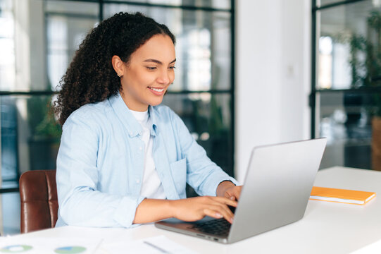 Successful motivated positive gorgeous multiracial curly haired woman in a shirt, employee of a company, software manager, sits at a work desk in the office, working on a project by laptop, smiles