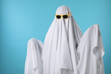 Stylish ghost. Person covered with white sheet in sunglasses on light blue background