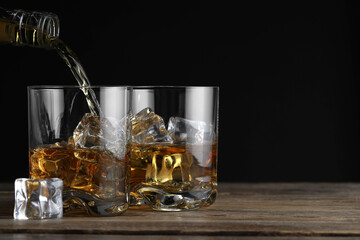 Pouring whiskey into glass with ice cubes at wooden table against black background, space for text